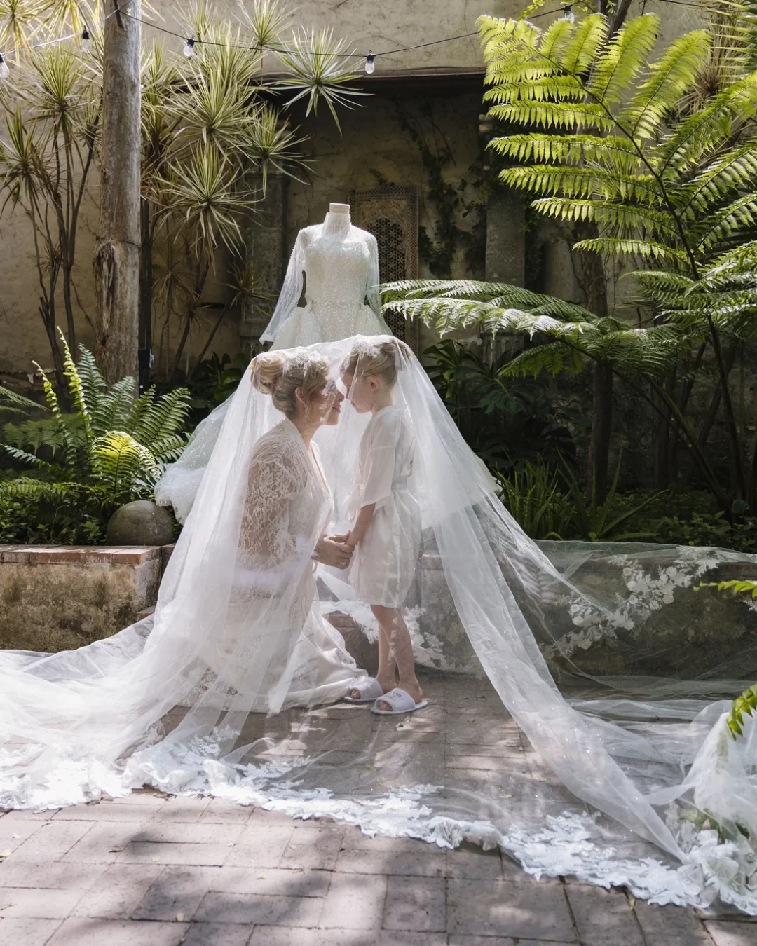 Retrato de novia y su hija con el velo extendido en el jardín